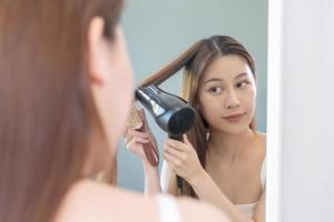 cabelo secador, feliz, ásia jovem mulher, menina olhando para dentro espelho refletir usando secador de cabelo para seco, sopro grandes Loiras em linha reta depois de chuveiro às lar. cabeleireiro, cabelo tratamento e beleza saúde Cuidado. foto