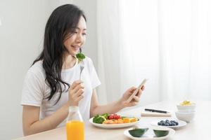 fazer dieta, ásia jovem mulher comendo, segurando garfo às brócolis, dieta plano nutrição com fresco legumes salada, apreciar refeição enquanto usando Smartphone. nutricionista do saudável, nutrição do peso perda. foto