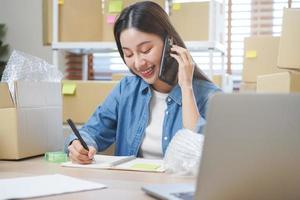 pequeno o negócio empreendedor sme, ásia jovem mulher proprietário embalagem produtos, verificação parcela caixa entrega, usando Smartphone receber ordem a partir de cliente, trabalhando às casa escritório. comerciante on-line, comércio eletrônico foto