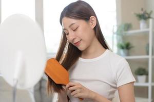 saúde cabelo Cuidado, beleza Maquiagem ásia mulher, menina segurando escova de cabelo e escovar, pentear dela grandes em linha reta cabelo olhando às reflexão dentro espelho dentro manhã rotina depois de salão tratamento, Penteado. foto