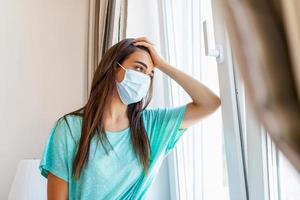 casa quarentena. caucasiano mulher sentado às janela dentro uma médico mascarar, olhando fora, quer para ir fora. proteção contra coronavírus infecção, pandemias, doença surtos e epidemias. foto