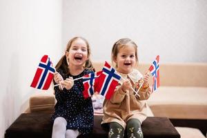 duas irmãs estão sentadas em um sofá em casa com bandeiras norueguesas nas mãos. noruega crianças meninas com bandeira. foto