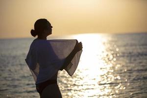 silhueta do uma mulher dentro uma branco toalha às pôr do sol em a mar. foto
