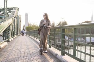 retrato do jovem o negócio mulher com a elétrico lambreta para trabalhos sobre ponte dentro moderno cidade fundo foto