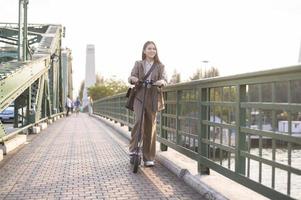 retrato do jovem o negócio mulher com a elétrico lambreta para trabalhos sobre ponte dentro moderno cidade fundo foto