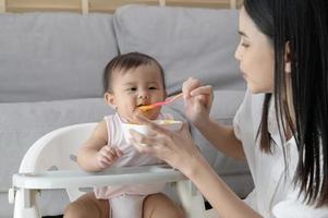 jovem mãe ajudando bebê comendo mistura Comida em bebê cadeira foto