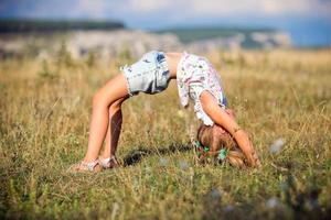 uma pequeno menina faz uma ponte, dobrar dela costas dentro natureza. uma flexível criança, fazendo ginástica exercícios. Esportes, alongamento, ioga, ativo estilo de vida conceitos foto