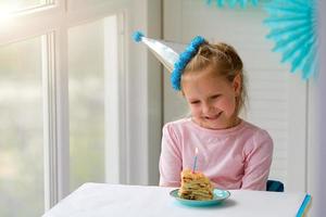 feliz pequeno menina senta dentro uma chapéu dentro frente do uma peça do bolo e uma vela. aniversário às lar. foto