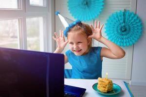 uma feliz pequeno garota, torto dentro frente do uma computador portátil comemora uma aniversário através da a Internet dentro quarentena tempo, isolamento voluntário, conectados aniversário foto