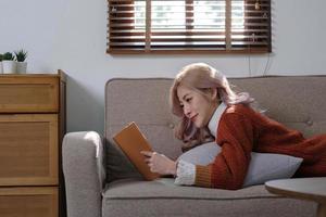 atraente jovem ásia mulher senta dentro a mínimo e confortável vivo quarto desfrutando lendo uma livro. foto