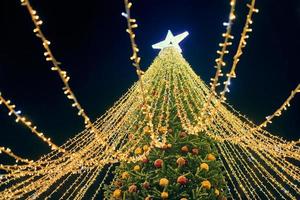 Natal árvore com amarelo guirlandas, decorativo lâmpadas e grande branco Estrela chapéu de coco às noite azul céu foto