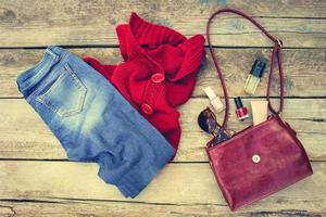 mulheres outono roupas e acessórios vermelho suéter, jeans, Bolsa, miçangas, oculos de sol e cosméticos em de madeira fundo. tonificado imagem. foto