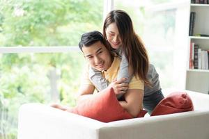 feliz casal sentado em a sofá e ser uma mulher abraçando dela namorado com amor dentro a vivo quarto e sorriso. conceito do romântico em namorados dia. proposta e casamento foto