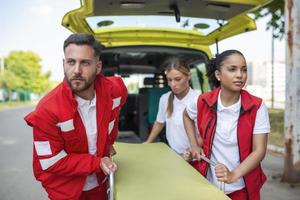 jovem paramédicos comovente ambulância maca a partir de carro dentro uma pressa. paramédicos dentro uniforme levando maca Fora a ambulância carro foto