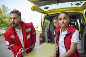 jovem paramédicos comovente ambulância maca a partir de carro. paramédicos dentro uniforme levando maca Fora a ambulância carro foto