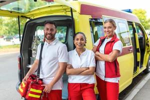 três multirracial paramédicos em pé dentro frente do ambulância veículo, carregando portátil equipamento foto