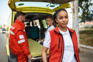 jovem fêmea africano americano paramédico em pé traseiro do a ambulância. paramédicos de a ambulância. dois paramédicos levando Fora maca a partir de ambulância foto