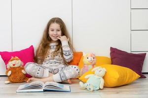 menina dentro pijamas senta em a chão entre colorida almofadas e a aberto livro e risos alegremente foto