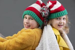 lindo pequeno menina e Garoto dentro tricotado Natal chapéus estão sentado com seus costas para cada de outros e sorridente em uma cinzento fundo. feliz Natal crianças. foto