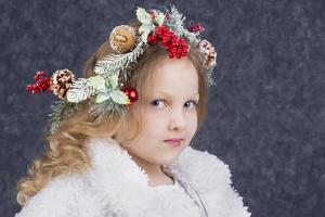 lindo pequeno Loiras menina com uma Natal guirlanda em dela cabeça em uma cinzento fundo. Natal criança. foto