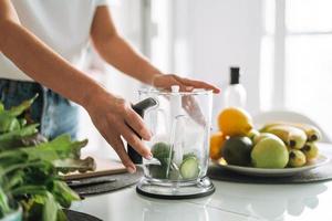 jovem fino mulher dentro branco camiseta e azul jeans cozinhando verde batido saudável Comida dentro cozinha às casa foto