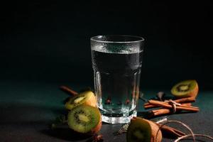 uma vidro do frio água em uma verde fundo. decorado com fatia kiwi, canela Gravetos, badyan, hortelã folhas. cópia de espaço foto