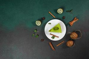 caseiro bolo de queijo com kiwi dentro uma branco prato em uma verde fundo. decorado com fatia do kiwi, canela bastão e folhas hortelã. topo Visão foto