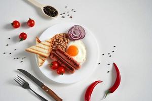 tradicional Inglês café da manhã. prato com frito ovo, dois frito salsichas, grelhado pão brinde, enlatado feijões, azul cebolas e cereja tomates em uma branco fundo. cópia de espaço foto