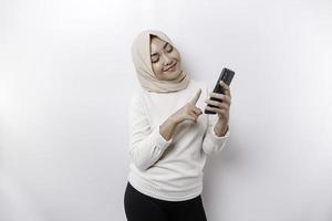 uma feliz ásia muçulmano mulher vestindo uma lenço de cabeça, segurando dela telefone, isolado de branco fundo foto