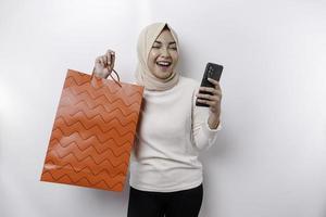 retrato ásia muçulmano mulher em pé animado segurando a conectados compras saco e dela Smartphone, estúdio tiro isolado em branco fundo foto