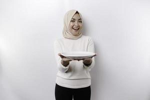 uma sorridente ásia muçulmano mulher é jejum e com fome e segurando e apontando para uma prato foto