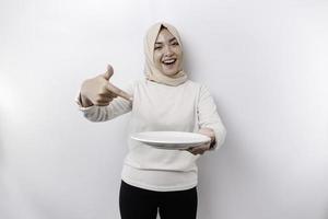 uma sorridente ásia muçulmano mulher é jejum e com fome e segurando e apontando para uma prato foto