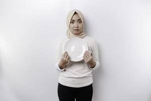 a ásia muçulmano mulher é jejum e com fome e segurando uma prato enquanto olhando a parte, de lado pensando sobre o que para comer foto