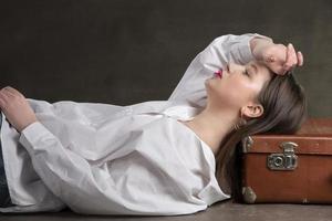 uma lindo menina mentiras em a velho mala de viagem em uma cinzento fundo. foto