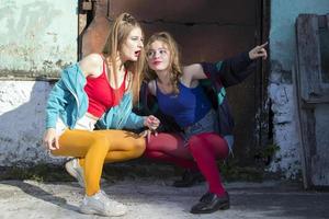 dois engraçado meninas dentro brilhante roupas dentro a estilo do a anos noventa estão sentado contra a fundo do uma oxidado porta. russo Vila garotas. foto