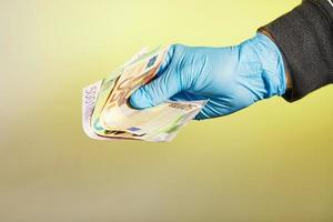 a mão de um homem com uma luva médica azul segura dinheiro na forma de notas de euro em um fundo amarelo foto