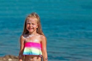 uma lindo pequeno menina sorrisos dentro uma roupa de banho em pé de a mar com protetor solar manchado em dela face. foto