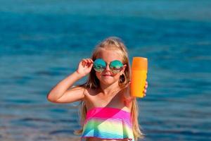 lindo pequeno menina dentro uma roupa de banho e oculos de sol carrinhos de a mar e detém uma protetor solar dentro dela mãos. foto