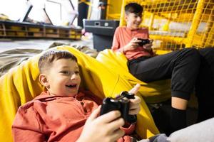 dois irmãos jogando vídeo jogos console, sentado em amarelo pufe dentro crianças jogar Centro. foto