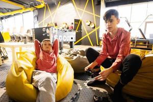 dois irmãos jogando vídeo jogos console, sentado em amarelo pufe dentro crianças jogar Centro. foto