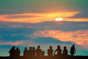 silhueta pessoas sentado em a topo montanha e pôr do sol em a colorida céu laranja nuvem e pássaros vôo foto