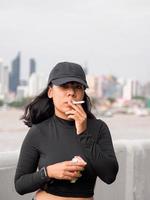 retrato mulher menina adolescente jovem Ásia 1 pessoa vestindo uma chapéu e grandes Preto cabelo vestindo uma Preto camisa mão aguarde fumar cigarro branco cor em pé ao ar livre de a parede ao ar livre foto