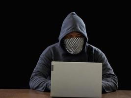 hacker espião homem uma pessoa com capuz preto sentado em uma mesa olhando computador laptop usado segurança de ataque de senha de login para circular dados digitais no sistema de rede da internet, fundo escuro noturno. foto