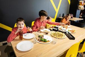 quatro crianças comendo italiano pizza dentro pizzaria. crianças comer às cafeteria. Garoto beber suco. foto