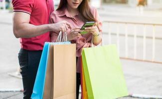 casal jovem feliz de compradores caminhando na rua comercial em direção e segurando sacolas coloridas na mão e usam um smartphone para promoção de cheques. conceito de venda e compras de sexta-feira negra foto