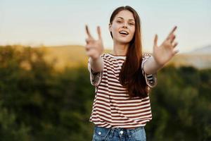 uma mulher sorrisos e puxa dela mãos para a Câmera fechar-se dentro natureza com uma Visão do a montanhas. feliz viagem estilo de vida Segue mim foto