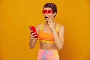 mulher blogueiro surpresa dentro incomum milenar óculos levando selfies em dela telefone esportivo brilhantemente colori roupas contra a laranja estúdio fundo, livre espaço foto