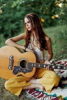 uma menina dentro roupas ecológicas hippie sentado com uma guitarra e olhando às uma pôr do sol dentro a verão foto