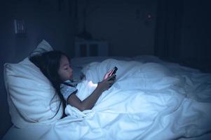 ásia criança jogando jogos em Smartphone dentro a cama às noite, o menina viciado social meios de comunicação foto