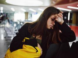 chateado mulher com amarelo mochila senta às a aeroporto enquanto esperando para uma voar foto
